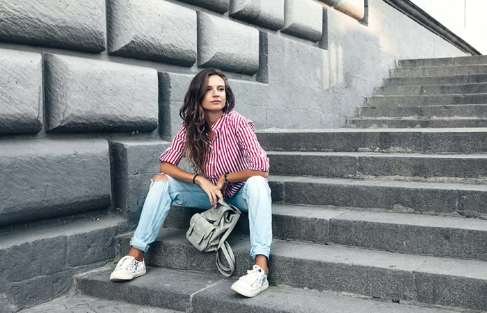 Model wearing a striped red shirt, ripped boyfriend jeans, white sneakers, and gray backpack posing in the stairs